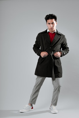 A man is standing confidently with his hands on the belt of a dark trench coat. He is wearing a red sweater, a white shirt underneath, gray plaid trousers, and casual sneakers. The background is a neutral gray, highlighting his fashion-forward look.