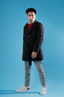 A person stands confidently against a blue background, wearing a black coat over a red shirt, gray pants, and white sneakers with red accents.