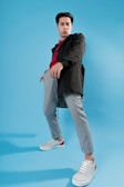 A man poses confidently against a bright blue background, wearing a black jacket, red sweater, and gray plaid pants. He stands with legs apart, gazing directly ahead with a bold expression. His outfit is completed with white sneakers that have red accents.