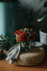 A bouquet of vibrant red flowers and green foliage is arranged in a glass vase. This vase is placed on a woven straw mat, accompanied by a delicate, patterned cloth. Surrounding the arrangement are potted plants and a tall teal planter on a wooden floor, creating a cozy and natural setting.