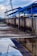 A rooftop setup with blue solar panels mounted on a metal frame, reflecting in a puddle of water on a concrete surface. The scene includes a cloudy sky in the background and additional pipes running along the ledge.