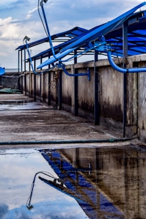 A rooftop setup with blue solar panels mounted on a metal frame, reflecting in a puddle of water on a concrete surface. The scene includes a cloudy sky in the background and additional pipes running along the ledge.
