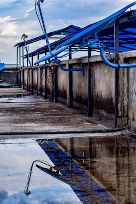 A rooftop setup with blue solar panels mounted on a metal frame, reflecting in a puddle of water on a concrete surface. The scene includes a cloudy sky in the background and additional pipes running along the ledge.