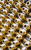 Numerous metallic brass fittings are arranged in a pattern, showcasing their shiny and reflective surfaces. The fittings have a hexagonal shape with threaded ends, indicating they are likely used in plumbing or machinery. The close-up angle emphasizes their geometric forms and metallic luster.