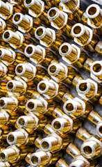Numerous metallic brass fittings are arranged in a pattern, showcasing their shiny and reflective surfaces. The fittings have a hexagonal shape with threaded ends, indicating they are likely used in plumbing or machinery. The close-up angle emphasizes their geometric forms and metallic luster.