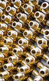 Numerous metallic brass fittings are arranged in a pattern, showcasing their shiny and reflective surfaces. The fittings have a hexagonal shape with threaded ends, indicating they are likely used in plumbing or machinery. The close-up angle emphasizes their geometric forms and metallic luster.