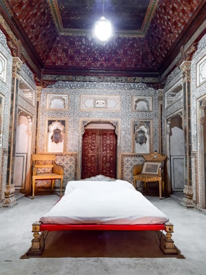 The image depicts an ornately decorated room with intricate patterns on the walls and ceiling. There is a bed with white linens centered in the room, flanked by traditional carved wooden chairs. The room has a historical or palace-like ambiance with rich detailing in the panel work and a chandelier hanging from the ceiling.