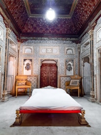 The image depicts an ornately decorated room with intricate patterns on the walls and ceiling. There is a bed with white linens centered in the room, flanked by traditional carved wooden chairs. The room has a historical or palace-like ambiance with rich detailing in the panel work and a chandelier hanging from the ceiling.