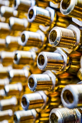 A close-up view of several threaded cylindrical metal objects, possibly bolts or connectors, arranged in a pattern. The metal surfaces have a shiny, polished appearance with golden and silver tones. The image has a shallow depth of field, creating a blurred effect in the background.
