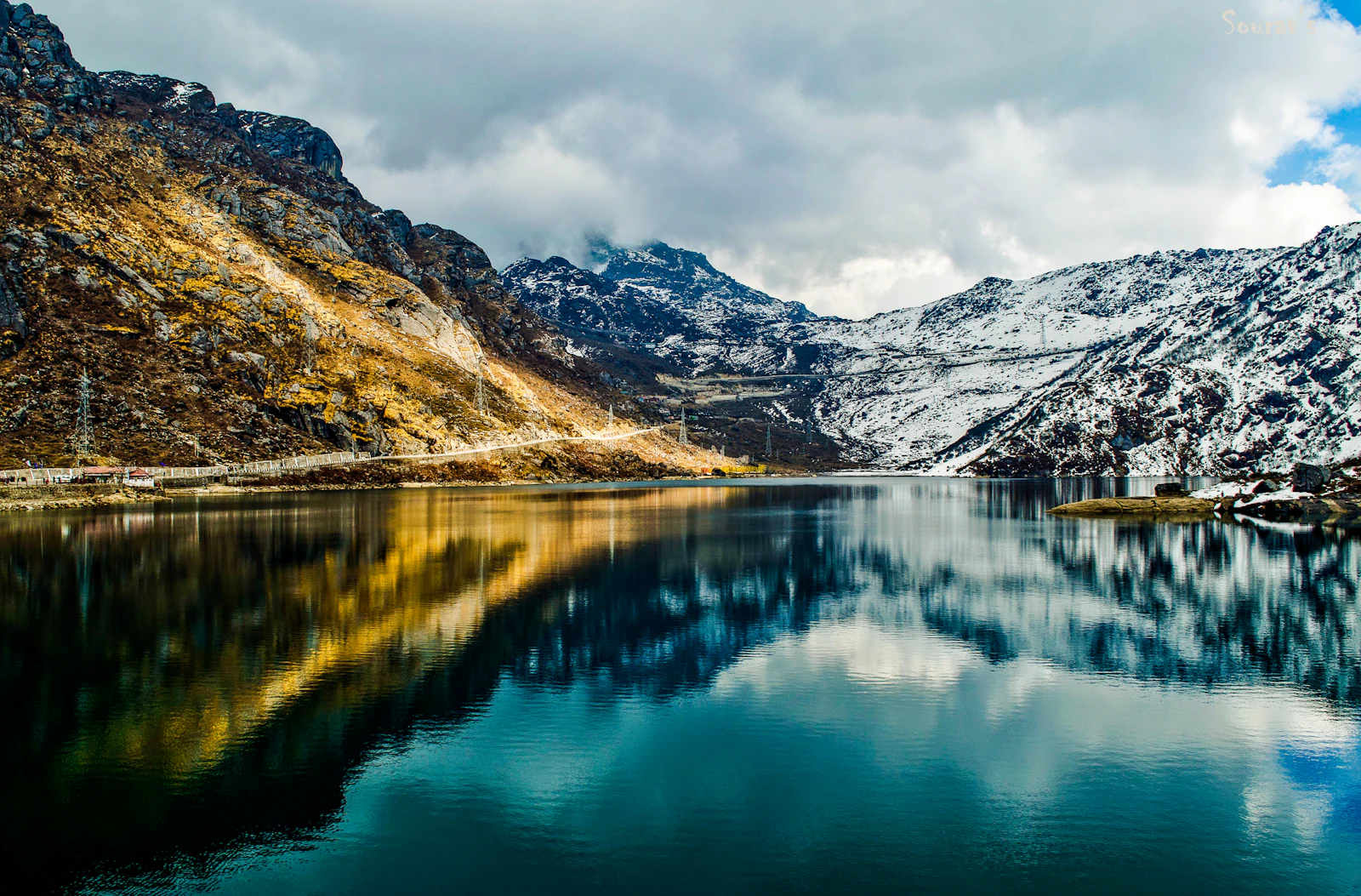 Tsomgo Lake in East Sikkim mirroring snow dusted peaks and a golden autumn slope, the Nathu La road traced along the far shore