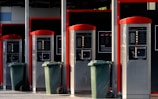 A row of four self-service washing machine controllers with red accents, each equipped with control panels and associated settings. In front of each station, there is a green waste bin on wheels. The area appears well-maintained and organized.