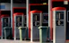 A row of four self-service washing machine controllers with red accents, each equipped with control panels and associated settings. In front of each station, there is a green waste bin on wheels. The area appears well-maintained and organized.