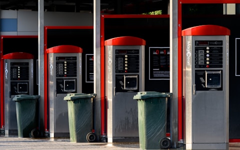 A row of four self-service washing machine controllers with red accents, each equipped with control panels and associated settings. In front of each station, there is a green waste bin on wheels. The area appears well-maintained and organized.