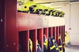 A row of red lockers is seen with yellow safety helmets placed on top of them. Below the helmets, fire service uniforms and jackets with reflective stripes are hanging inside the open lockers. The background includes a white wall with some indistinct elements.