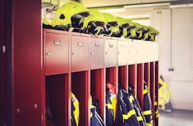 A row of red lockers is seen with yellow safety helmets placed on top of them. Below the helmets, fire service uniforms and jackets with reflective stripes are hanging inside the open lockers. The background includes a white wall with some indistinct elements.