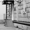 A monochrome image of a building with a textured exterior and a small staircase leading to a doorway. A man is leaning out of a window, holding a mobile phone next to his ear. The building has a sign with Hebrew text.