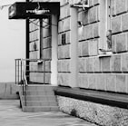 A monochrome image of a building with a textured exterior and a small staircase leading to a doorway. A man is leaning out of a window, holding a mobile phone next to his ear. The building has a sign with Hebrew text.