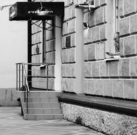 A monochrome image of a building with a textured exterior and a small staircase leading to a doorway. A man is leaning out of a window, holding a mobile phone next to his ear. The building has a sign with Hebrew text.