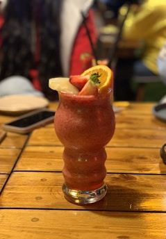A vibrantly colored fruit smoothie is presented in a uniquely shaped glass, garnished with a slice of lemon, an orange, and a strawberry on a wooden table. The blurred background suggests a lively social setting.