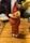 A vibrantly colored fruit smoothie is presented in a uniquely shaped glass, garnished with a slice of lemon, an orange, and a strawberry on a wooden table. The blurred background suggests a lively social setting.