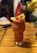 A vibrantly colored fruit smoothie is presented in a uniquely shaped glass, garnished with a slice of lemon, an orange, and a strawberry on a wooden table. The blurred background suggests a lively social setting.