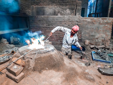 A person wearing a white outfit and a pink headscarf is working with fire in a rustic, brick-walled room. They are tending to a flame with a long metal tool, surrounded by mounds of dirt and metal castings. Various tools and materials are scattered around, highlighting an industrial or artisanal metalwork setting.