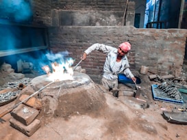 A person wearing a white outfit and a pink headscarf is working with fire in a rustic, brick-walled room. They are tending to a flame with a long metal tool, surrounded by mounds of dirt and metal castings. Various tools and materials are scattered around, highlighting an industrial or artisanal metalwork setting.