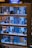 A model of a multi-floor office building with miniature figures depicting people engaging in various office activities. The building has several floors, each with different setups and figures, such as desks, office equipment, and people interacting. The top of the building has a sign that reads 'Transportation'. The floors appear to be lit, showcasing an interior view of an office environment.