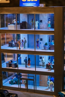 A model of a multi-floor office building with miniature figures depicting people engaging in various office activities. The building has several floors, each with different setups and figures, such as desks, office equipment, and people interacting. The top of the building has a sign that reads 'Transportation'. The floors appear to be lit, showcasing an interior view of an office environment.
