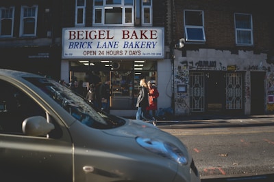 A street scene featuring a bakery with a bright sign reading 'Beigel Bake Brick Lane Bakery' that is open 24 hours a day. People are seen entering and exiting the shop, with one individual dressed in a red coat visible on the sidewalk. A car is parked in the foreground, partially obscuring the view of the shop. The building exterior is covered with graffiti.