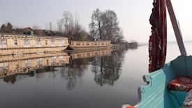 A serene body of water reflects a row of traditional houseboats and bare trees, creating a symmetrical image. The foreground includes part of a boat with decorative elements and a patterned cushion, leading into the tranquil scene.