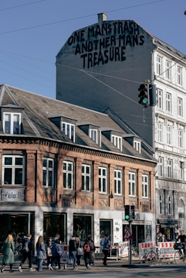 A multi-story urban building with a mural featuring the phrase 'One man's trash, another man's treasure' on the upper wall. The building has a brick facade with large windows displaying bicycles. Pedestrians are crossing the street near traffic lights, and red and white barricades are visible.