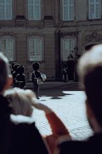 Several guards in traditional uniforms stand in formation on a cobblestone courtyard in front of a historic building with large windows. A few people in the foreground appear to be observing the scene.