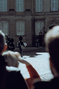 Several guards in traditional uniforms stand in formation on a cobblestone courtyard in front of a historic building with large windows. A few people in the foreground appear to be observing the scene.