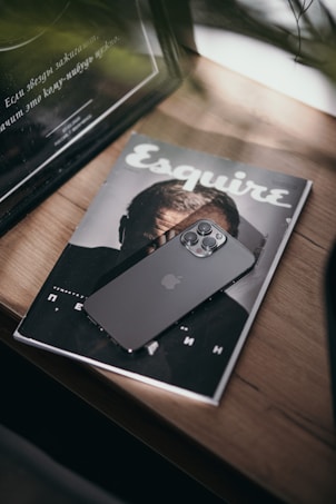 A sleek, modern smartphone with multiple camera lenses rests on the cover of a magazine. The magazine's cover shows a blurred image of a person. The scene is set on a wooden surface, with soft, natural lighting and some greenery in the foreground adding depth to the composition.