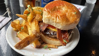 A juicy burger topped with crispy bacon, melted cheese, and a slice of tomato is served on a toasted bun. It is accompanied by a generous portion of thick, golden-brown fries on a white plate. Sprinkles of herbs and spices add a touch of color. The setting includes a dark reflective table surface and a glimpse of beverages in the background.