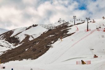Snow-covered mountain slopes with ski lifts running along the terrain. Red safety fences line the skiing paths. A partly cloudy sky provides a backdrop.