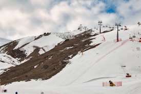 Snow-covered mountain slopes with ski lifts running along the terrain. Red safety fences line the skiing paths. A partly cloudy sky provides a backdrop.