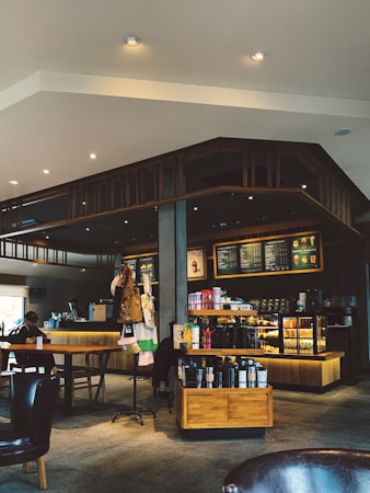 A cozy coffee shop interior featuring a wooden counter lined with a variety of coffee machines and bottled drinks. The menu board above the counter displays various coffee options. There are tables with chairs, and a person sits at one of them. The lighting is warm, enhancing the comfortable ambiance of the space.