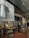 An upscale indoor cafe or bar setting with wooden and metallic accents. There are wooden tables with unique wireframe bases and leather chairs. A red patterned carpet adds color to the wooden flooring. On the wall, a large black-and-white mural features a man with headphones, dressed in a suit and tie. Next to it, a smaller art piece shows two abstract faces.