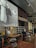 An upscale indoor cafe or bar setting with wooden and metallic accents. There are wooden tables with unique wireframe bases and leather chairs. A red patterned carpet adds color to the wooden flooring. On the wall, a large black-and-white mural features a man with headphones, dressed in a suit and tie. Next to it, a smaller art piece shows two abstract faces.