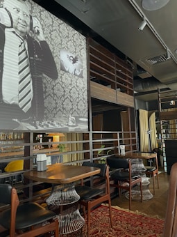 An upscale indoor cafe or bar setting with wooden and metallic accents. There are wooden tables with unique wireframe bases and leather chairs. A red patterned carpet adds color to the wooden flooring. On the wall, a large black-and-white mural features a man with headphones, dressed in a suit and tie. Next to it, a smaller art piece shows two abstract faces.