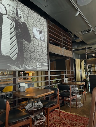 An upscale indoor cafe or bar setting with wooden and metallic accents. There are wooden tables with unique wireframe bases and leather chairs. A red patterned carpet adds color to the wooden flooring. On the wall, a large black-and-white mural features a man with headphones, dressed in a suit and tie. Next to it, a smaller art piece shows two abstract faces.