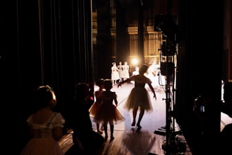 Several dancers in costumes are gathered backstage with some illuminated by stage lights in the background. The scene captures the anticipation and behind-the-scenes atmosphere of a performance, with figures in silhouette and bright stage lighting creating contrast.