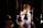 Several dancers in costumes are gathered backstage with some illuminated by stage lights in the background. The scene captures the anticipation and behind-the-scenes atmosphere of a performance, with figures in silhouette and bright stage lighting creating contrast.