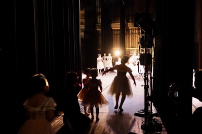 Several dancers in costumes are gathered backstage with some illuminated by stage lights in the background. The scene captures the anticipation and behind-the-scenes atmosphere of a performance, with figures in silhouette and bright stage lighting creating contrast.