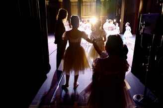 Dancers in elegant costumes stand backstage, illuminated by bright stage lights as they prepare to perform. The setting is a theater, with curtains partially open to reveal the stage. The atmosphere is one of anticipation and excitement.