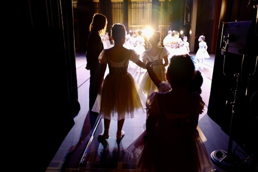 Dancers in elegant costumes stand backstage, illuminated by bright stage lights as they prepare to perform. The setting is a theater, with curtains partially open to reveal the stage. The atmosphere is one of anticipation and excitement.