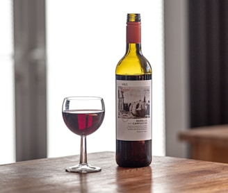 A bottle of red wine stands on a wooden table next to a filled wine glass. The label on the bottle displays text and an image in black and white. Bright light streams in from a window in the background, creating a cozy and relaxed atmosphere.