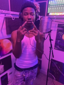 A person wearing headphones and a white tank top stands in a music studio. They are gesturing with their hands, positioned in front of a microphone with a pop filter. The room has purple lighting and is surrounded by soundproof panels and various artworks. A computer screen with audio mixing software is visible in the background.
